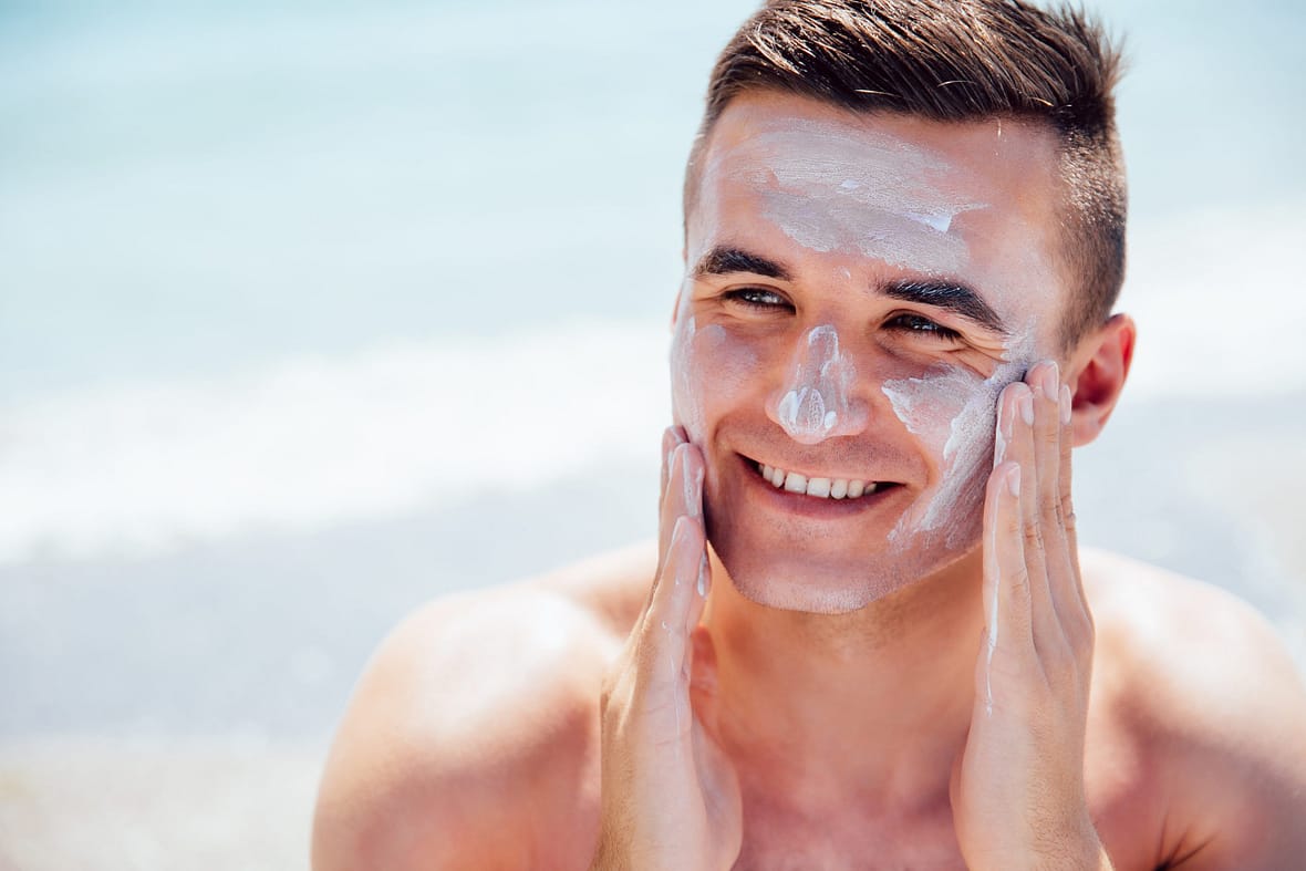 Smiling man putting tanning cream on his face, takes a sunbath on the beach. Healthcare.