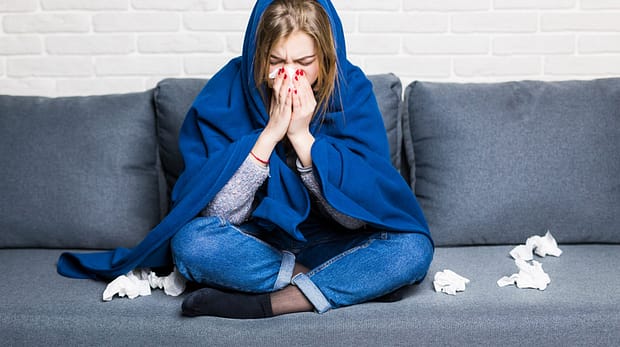 Sick woman with rheum and headache holding napkin, sitting on sofa with coverlet and pills at home