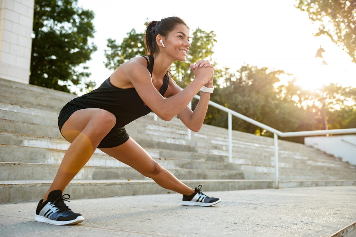 Portrait of a smiling fitness woman in earphones doing stretching exercises on stairs outdoors