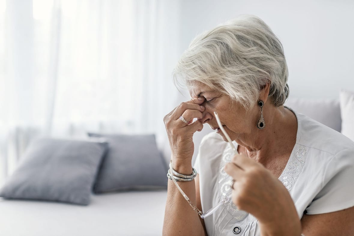 Mature woman sitting on a white sofa in a home touching her head with her hands while having a headache pain and feeling unwell. Senior woman with headache