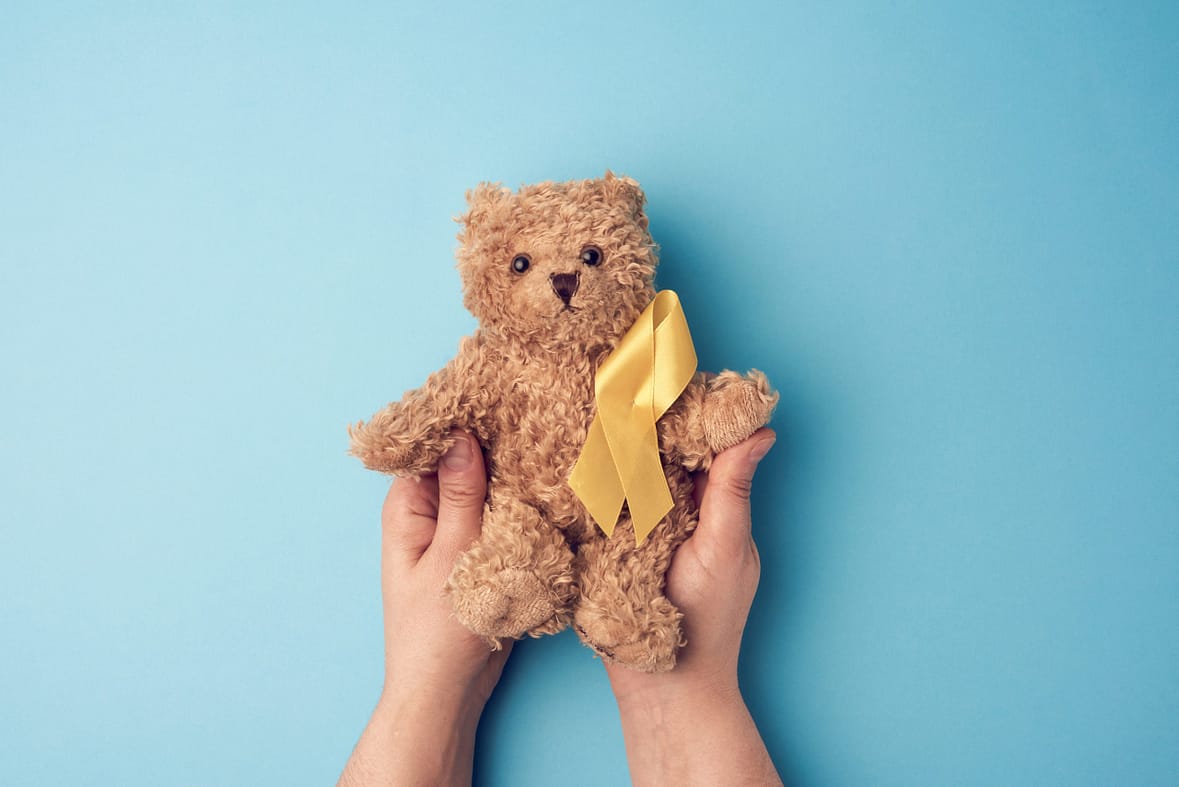 female hands hold a small teddy bear with a yellow ribbon folded in a loop on a blue background. concept of the fight against childhood cancer. problem of suicides