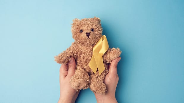 female hands hold a small teddy bear with a yellow ribbon folded in a loop on a blue background. concept of the fight against childhood cancer. problem of suicides